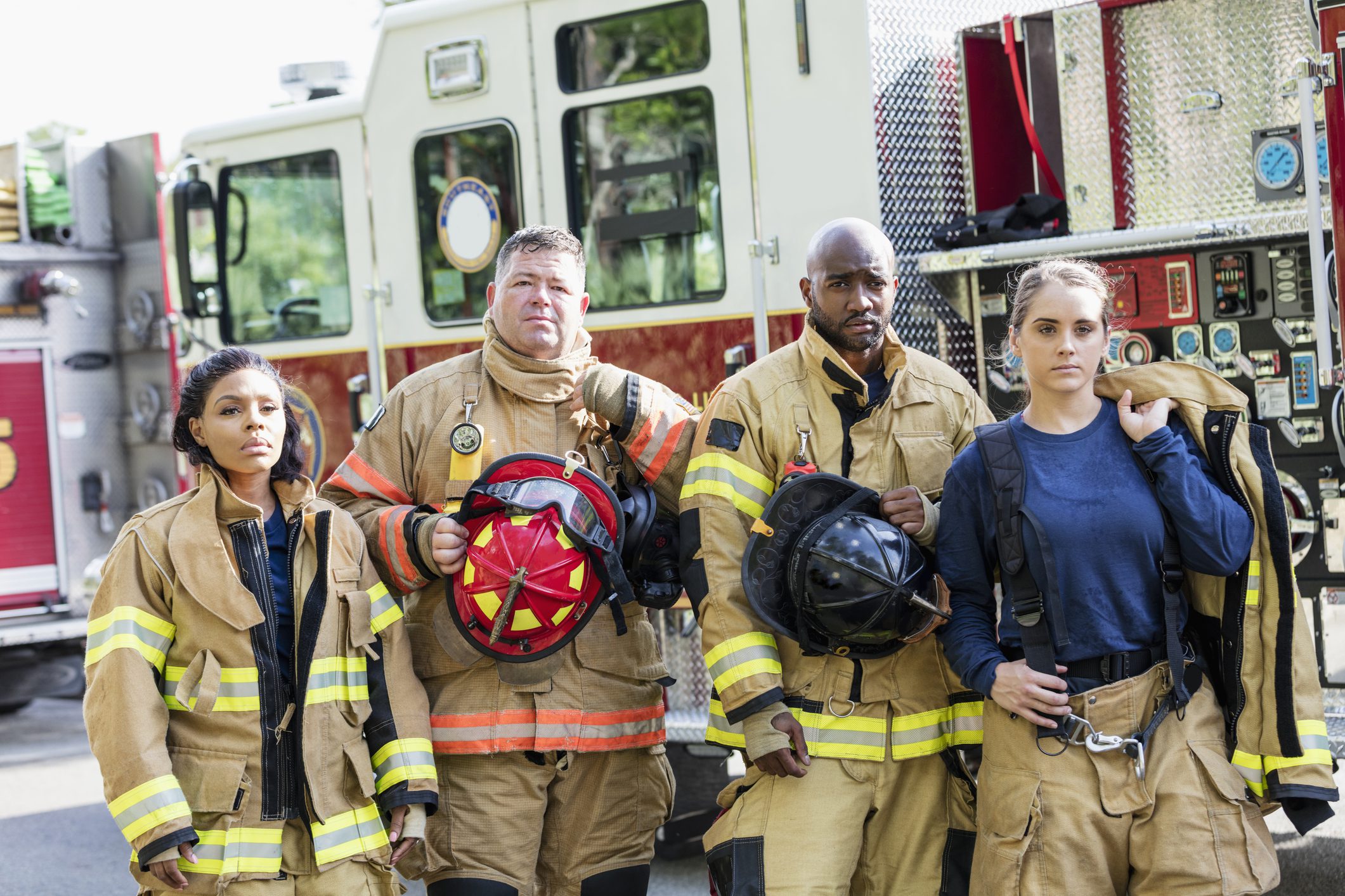 A multi-ethnic group of four firefighters standing together in front of fire engines, wearing fire protection suits and holding helmets. Two of them are women. They are looking at the camera with serious expressions.